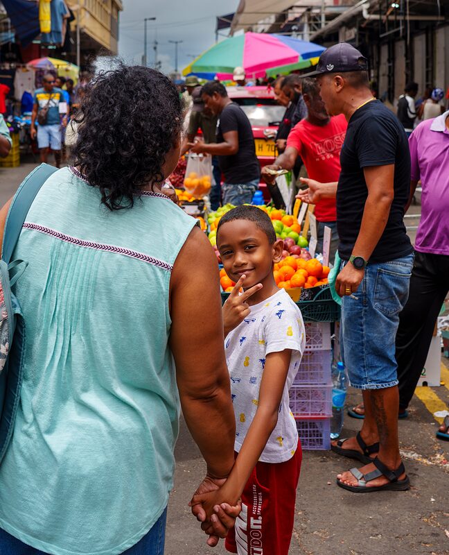 Jeune garçon dans le marché de Port-Louis, accompagné d'une adulte qui fait un signe amical au photographe. Île Maurice, 2026.